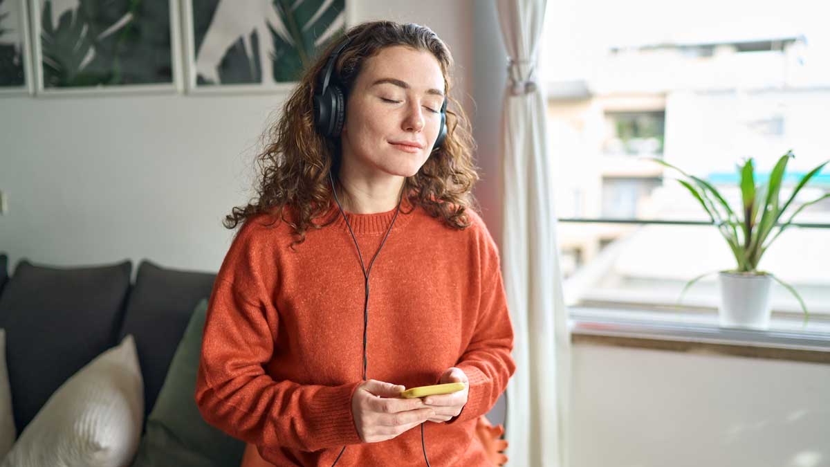 Mujer escuchando música con audífonos en su sala, sonriendo relajada.