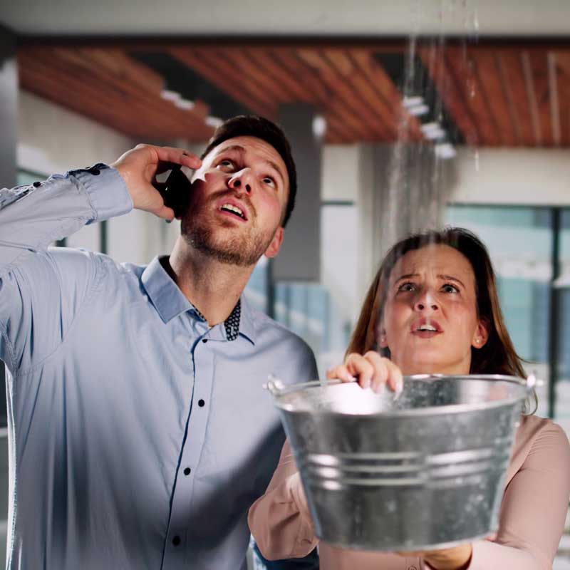 Pareja hombre-mujer dentro de casa con cubeta por gotera y daños por agua en casa.