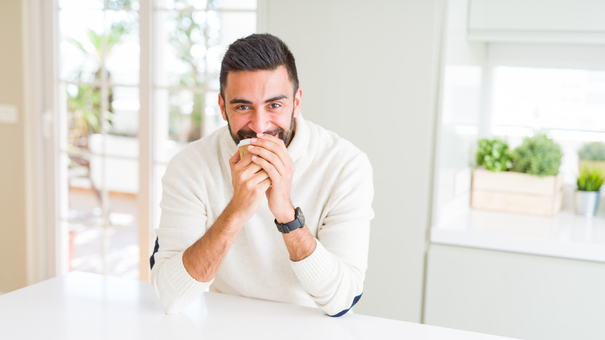 Hombre con barba, sonriendo en la cocina de su casa tomando café.