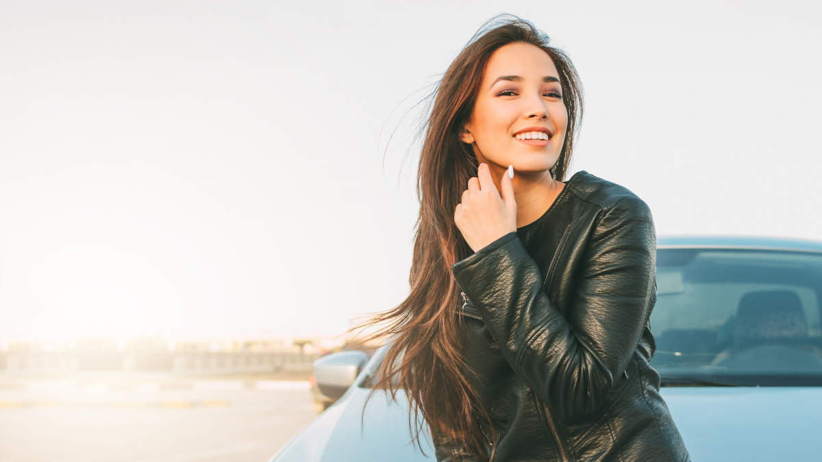 Mujer joven sonriendo frente de su automóvil en un estacionamiento al atardecer.