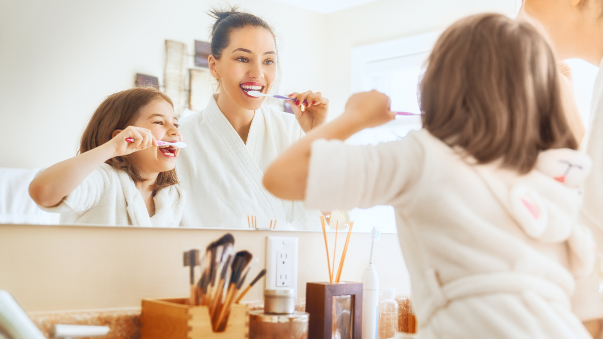[Madre e hija sonriendo frente al espejo sosteniendo un cepillo de dientes. Mujer joven preparándose para su rutina matutina de higiene bucal en el baño