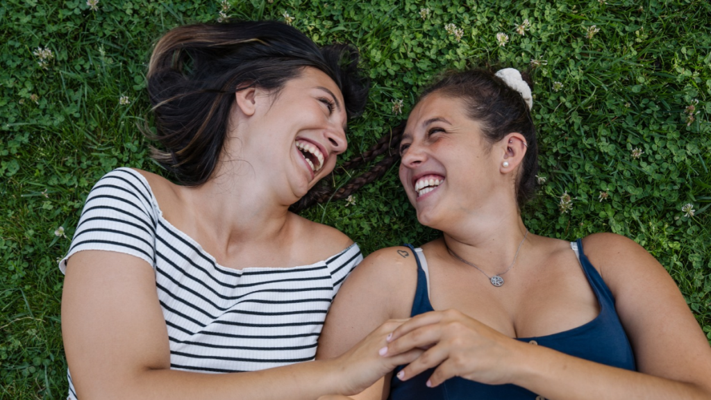 Dos mujeres sonriendo y tomadas de la mano recostadas sobre el césped, símbolo de apoyo, salud femenina y sororidad.