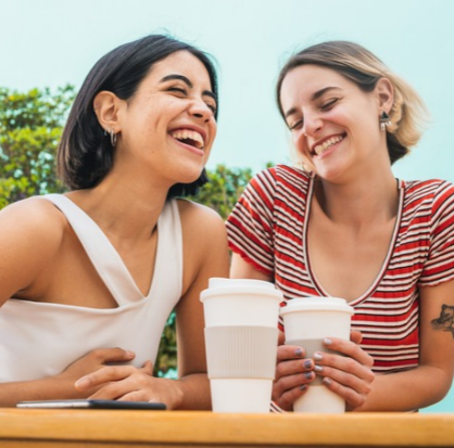 Dos mujeres compartiendo risas y café en una terraza, representando el valor de la amistad, el bienestar emocional y el apoyo entre mujeres.